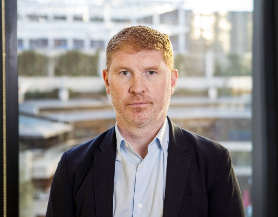 Man in shirt and black suit jacket standing in front of a window with office buildings in the background