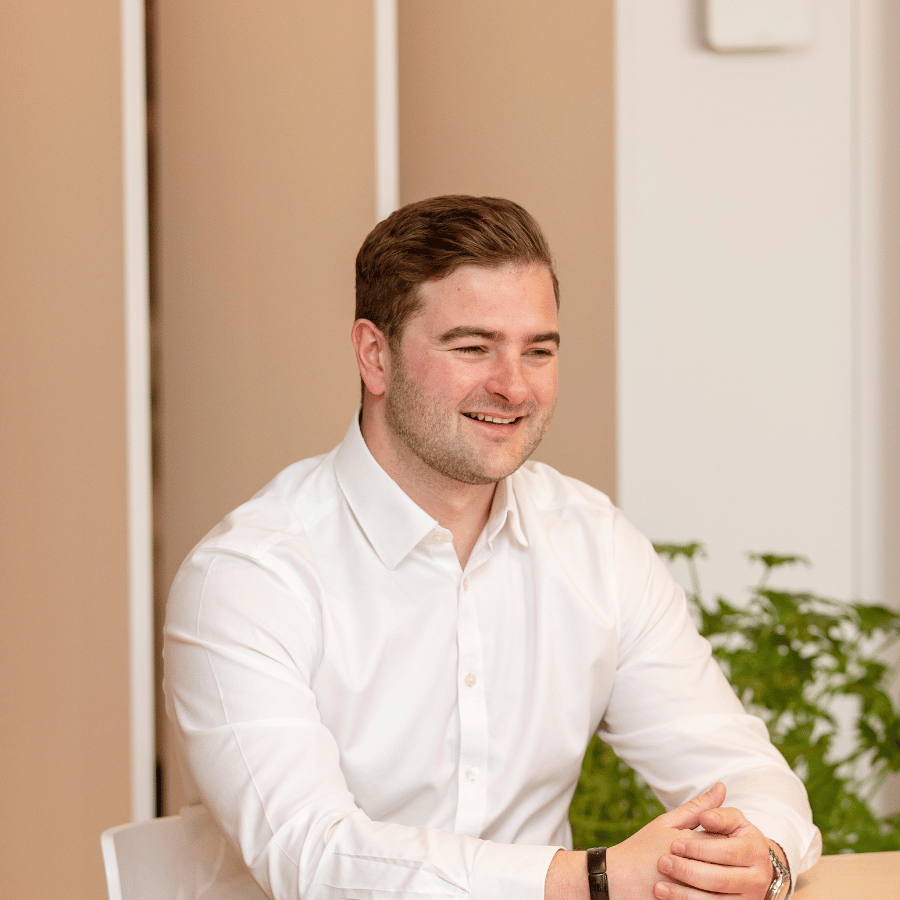 Man in a white shirt sitting on a chair at a table, with beige panels and a green plant in the background.