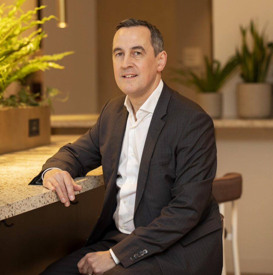 Man in white shirt and grey jacket leaning against a counter top in breakout space.