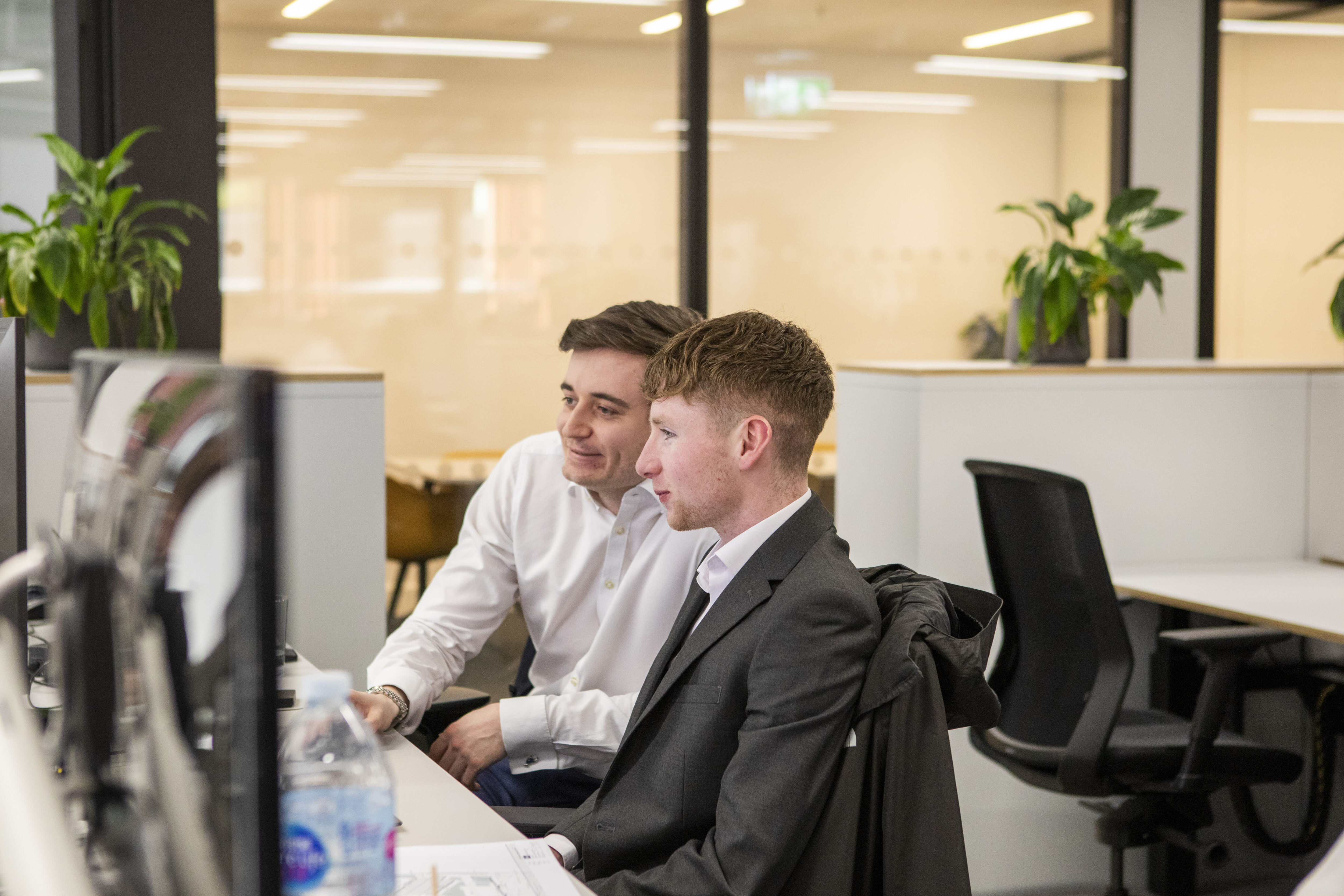 Two workers in a modern white office space looking at computer surrounded by green plants.