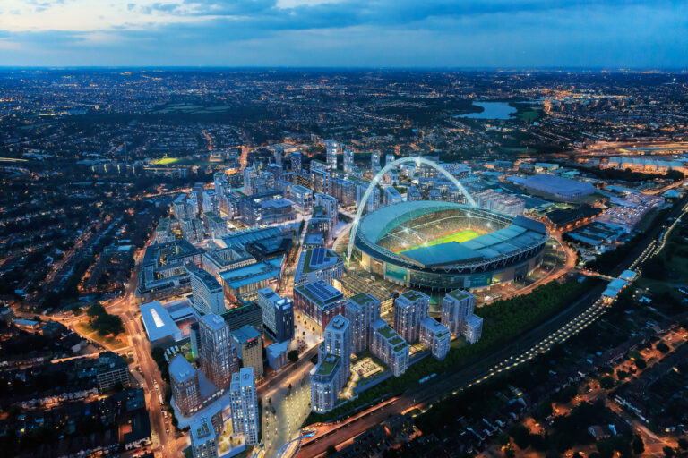 A sold out Wembley stadium surrounded by a large amount of modern high top and low top buildings with lights on at night.