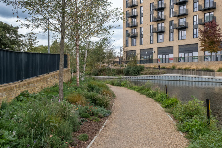 A park lined with lush greenery curving behind a tranquil pond. A modern brick building with balconies in the background.