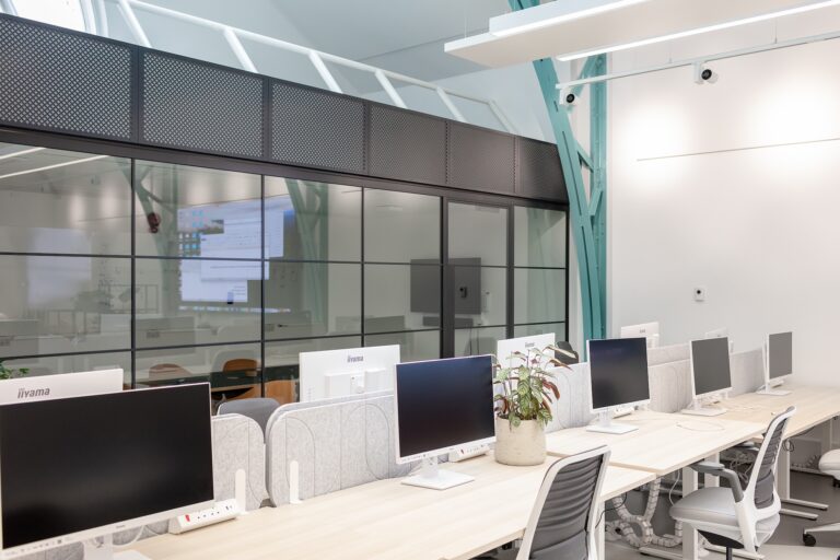 A room featuring a table surrounded by chairs, with a desk and computer setup against the wall. The space is designed as an office with various pieces of furniture, including an office chair, and includes a ceiling overhead.