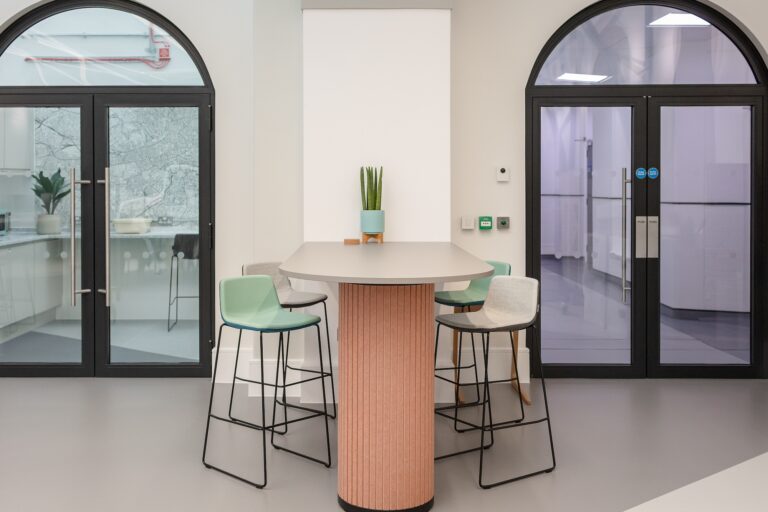 A wooden table surrounded by chairs in a cozy room, with a large mirror on the wall, a window letting in natural light, and decorative elements that create an inviting atmosphere.