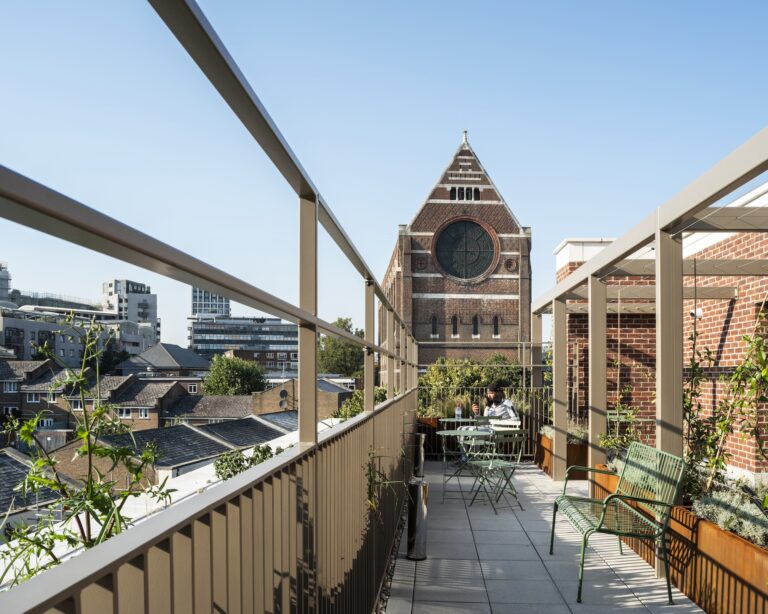 A balcony with a view of a clock tower and nearby building, surrounded by plants and flowerpots, set against a clear sky in an urban environment.