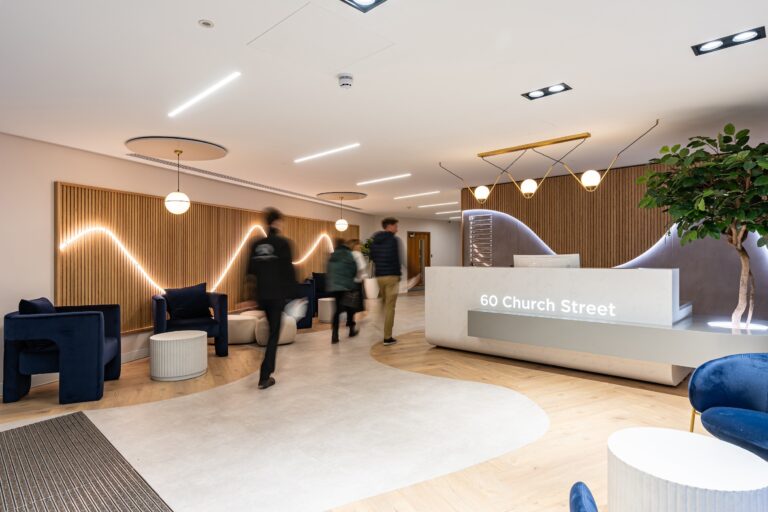 A group of people is gathered in a room featuring a modern interior design with a ceiling, walls, and flooring. There are various pieces of furniture including a studio couch and a loveseat arranged to create a comfortable sitting area, resembling a hotel lobby or waiting room.