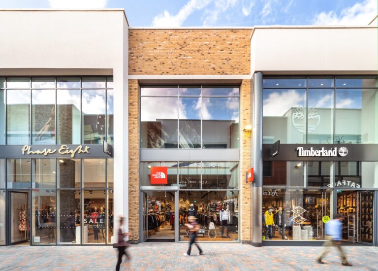 A commercial building with glass windows, labeled with signs for Phase Eight, Timberland, AITAS, and ALE. The structure is situated in a city environment with a blue sky and clouds overhead. People can be seen nearby, and the scene includes elements of a mall setting on the ground level.