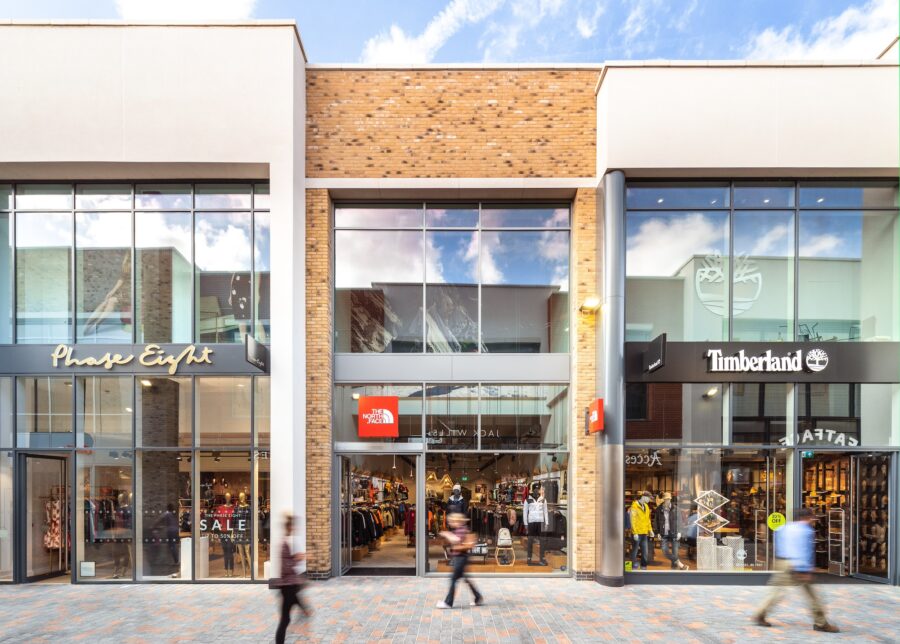 A commercial building with glass windows, labeled with signs for Phase Eight, Timberland, AITAS, and ALE. The structure is situated in a city environment with a blue sky and clouds overhead. People can be seen nearby, and the scene includes elements of a mall setting on the ground level.