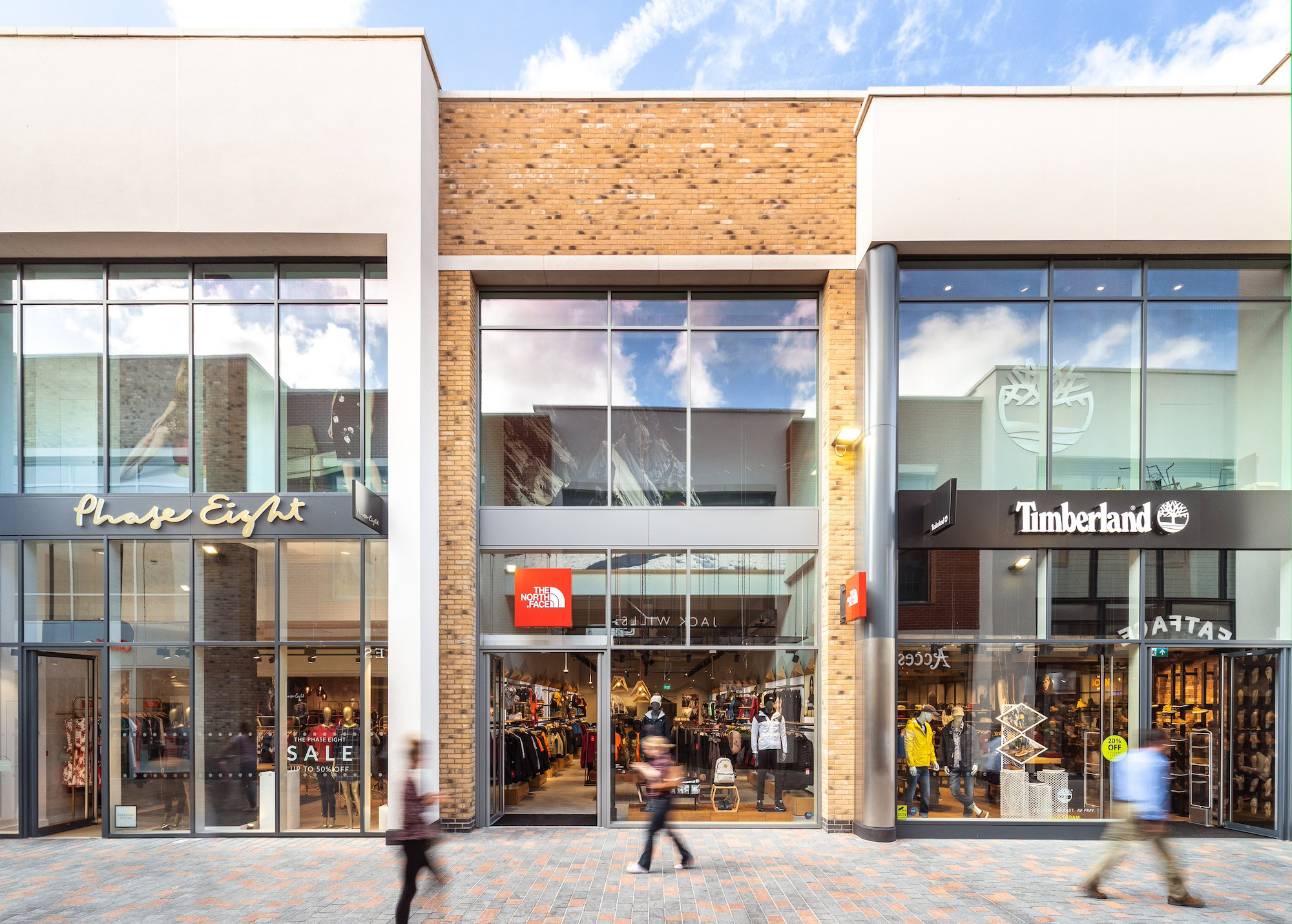 A commercial building with glass windows, labeled with signs for Phase Eight, Timberland, AITAS, and ALE. The structure is situated in a city environment with a blue sky and clouds overhead. People can be seen nearby, and the scene includes elements of a mall setting on the ground level.
