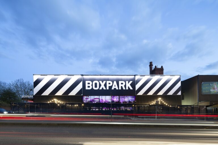 A building with a sign that reads "BOXPARK" displayed prominently. The setting includes a clear blue sky with some clouds overhead, surrounded by outdoor elements such as a road and street signage. The area appears to be vibrant and inviting for shoppers.