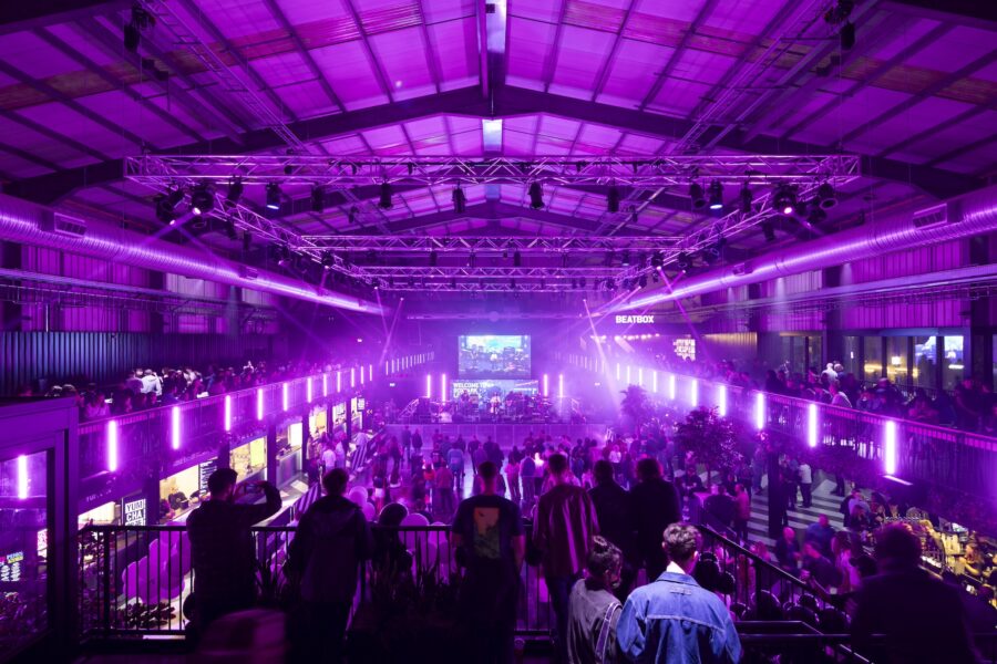 A group of people gathered in a dimly lit room, featuring a large screen and vibrant lights, reminiscent of a nightclub atmosphere.