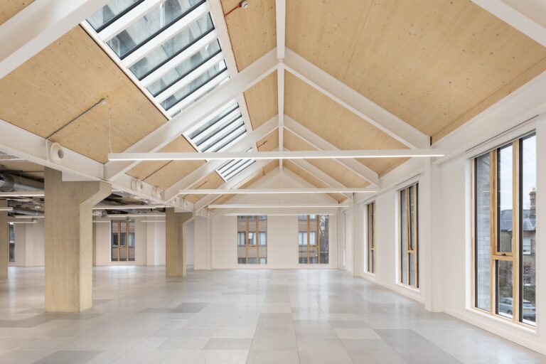 A large, bright white room featuring white walls, a ceiling with fixtures and beams, and symmetrical architectural design elements. The space includes a floor and windows that allow daylight to fill the room.