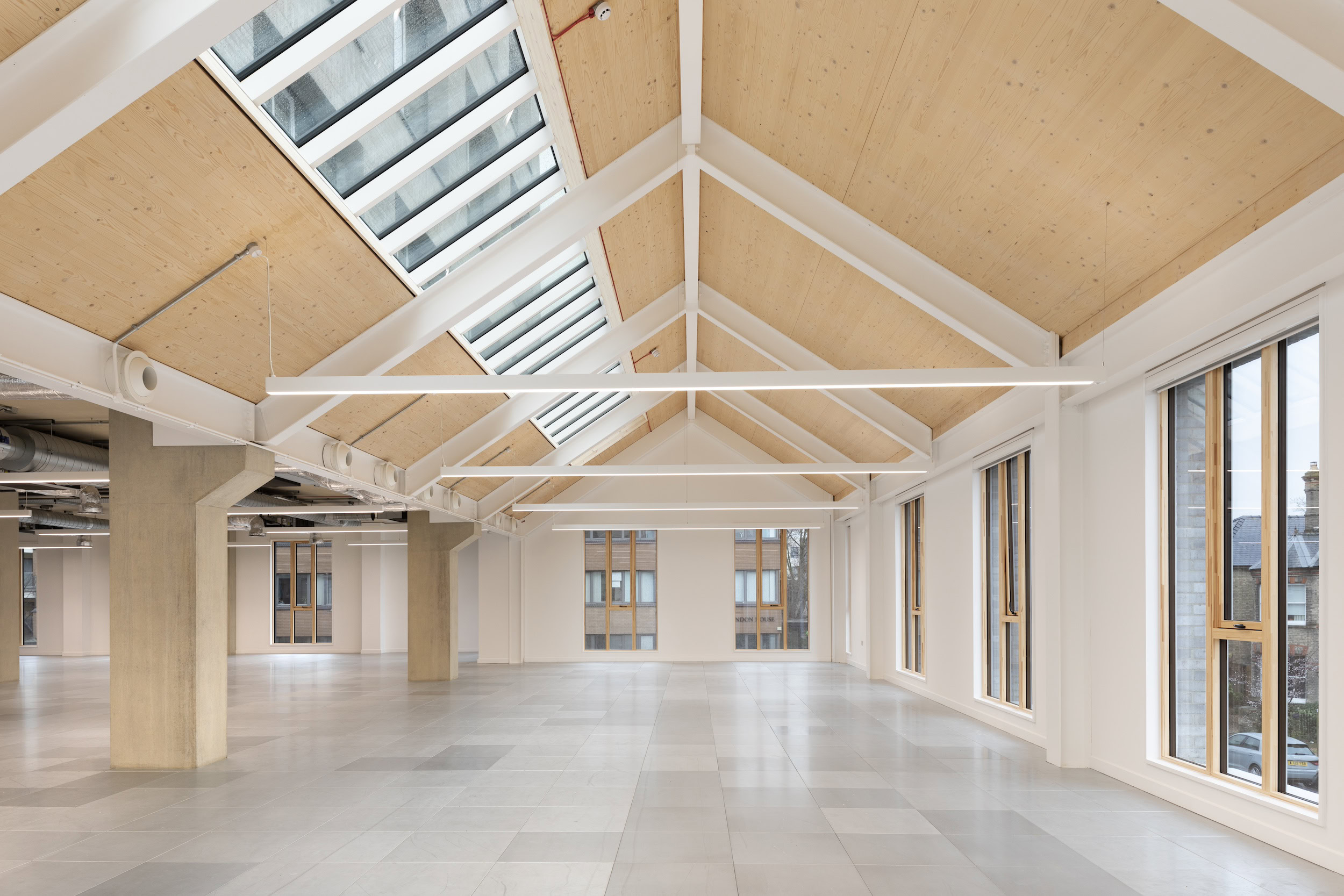 A large, bright white room featuring white walls, a ceiling with fixtures and beams, and symmetrical architectural design elements. The space includes a floor and windows that allow daylight to fill the room.
