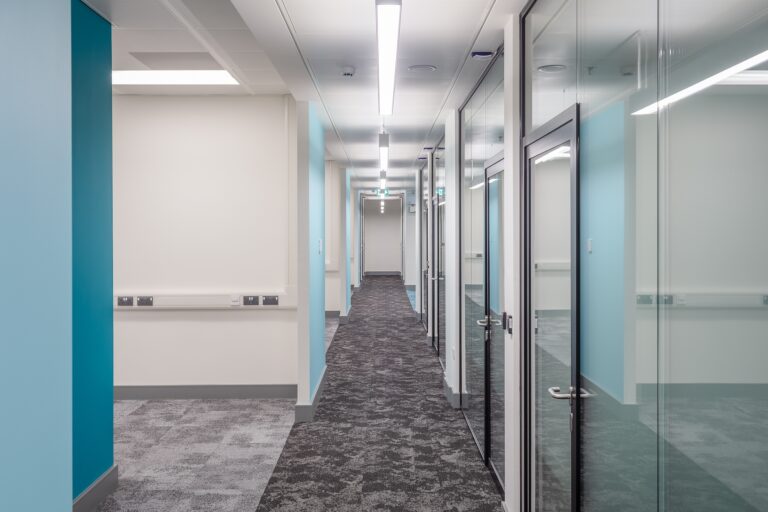 A hallway featuring glass doors, with walls and flooring designed for an indoor space. The interior design includes ceiling fixtures and a clean floor.