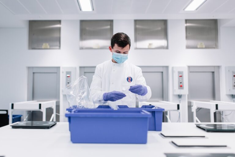 Placeholder A man wearing a mask and gloves stands indoors in a laboratory setting, surrounded by medical equipment and research instruments. He is dressed in a white coat, signaling his role as a scientist or researcher. The background features walls typical of a healthcare facility.