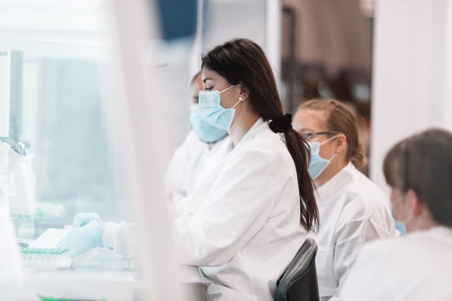 A woman wearing a white coat stands indoors in a laboratory setting. She is depicted as a healthcare provider, possibly a scientist or medical technologist, surrounded by various laboratory equipment and materials.