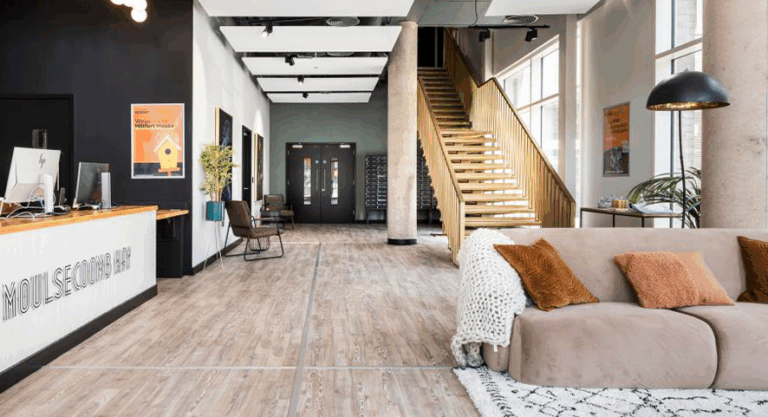 A room featuring a couch and a staircase, showcasing an indoor interior design with laminate flooring and wood accents. The space resembles a loft or penthouse apartment, designed for comfort and style.
