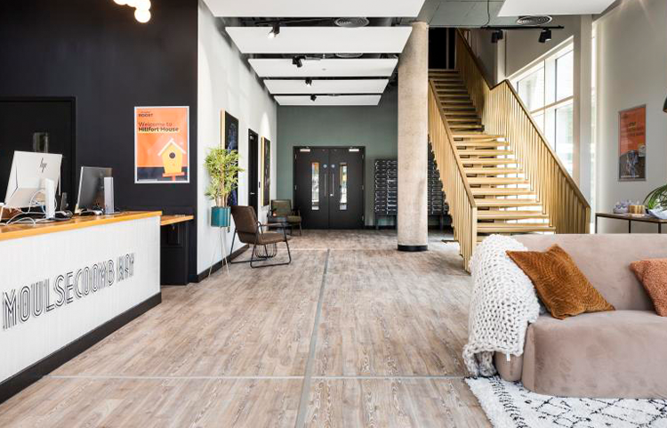 A room featuring a couch and a staircase, showcasing an indoor interior design with laminate flooring and wood accents. The space resembles a loft or penthouse apartment, designed for comfort and style.