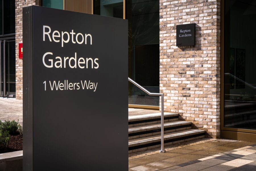 A sign on a building displaying the text "Repton Gardens 1 Wellers Way." The sign is mounted on a brick wall with a window and door nearby, and there are stairs leading up to the entrance. The setting is an outdoor city environment with a sidewalk.