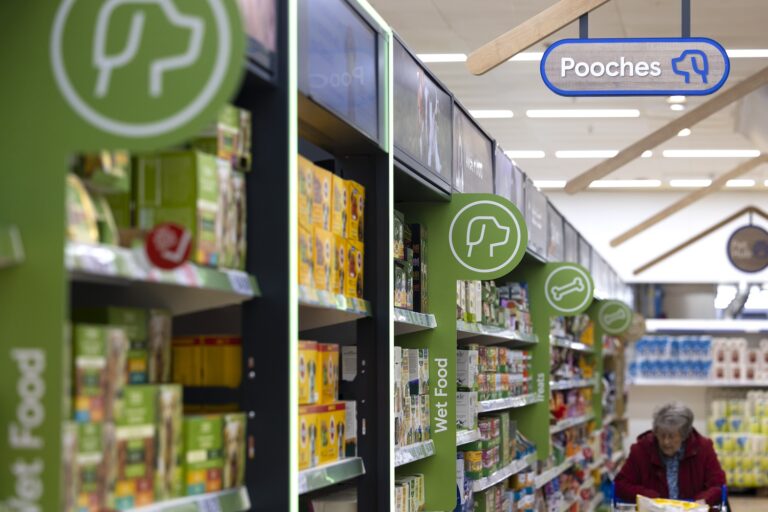 Inside Tesco's store in Cheshunt, displaying a person walking down a dog food isle in a modern complex. Green signs, with dog logos and a large amount of products.