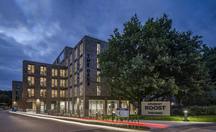Modern student accommodation building named "The Oaks" at dusk, with illuminated windows, signage for "Student Roost," and surrounding trees.