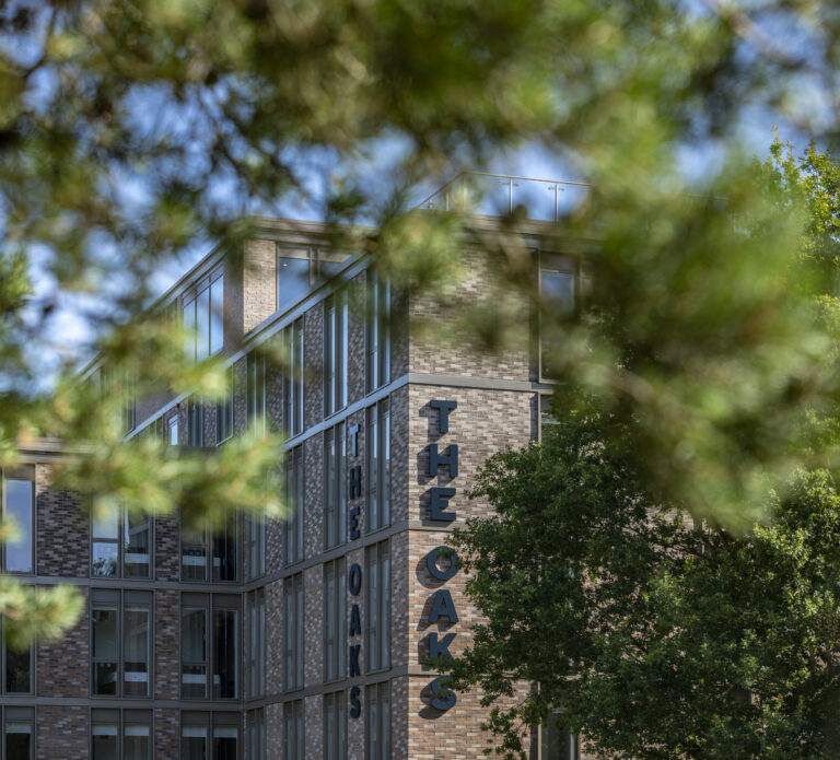 View of "The Oaks" student accommodation building through tree branches on a sunny day.