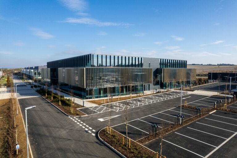 A large building stands in the background with a road leading up to it, under a sky filled with clouds. The area features parking spaces nearby.
