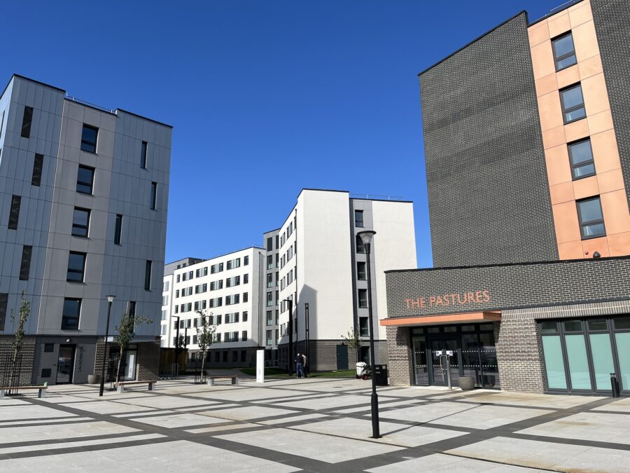 A view of a group of buildings surrounding a courtyard in an urban setting, with a clear sky above. The structures are commercial and mixed-use, featuring modern architecture and large facades. A road runs alongside the buildings in the downtown city area.