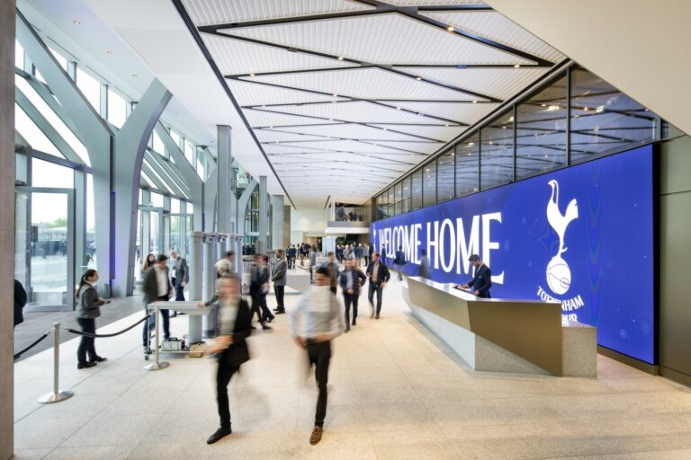 A spacious lobby wiht a blue digital screen reading "Welcome Home" and a logo. A diverse group of people walking together inside a building, with visible footwear and clothing. The interior features a ceiling and floor design, along with artistic elements in the surroundings.