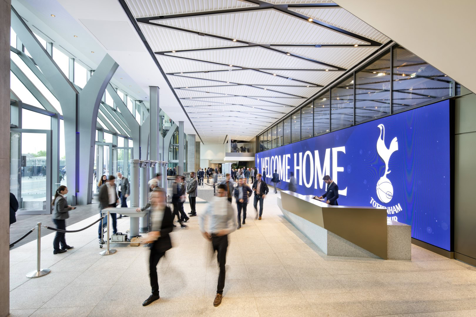 A spacious lobby wiht a blue digital screen reading "Welcome Home" and a logo. A diverse group of people walking together inside a building, with visible footwear and clothing. The interior features a ceiling and floor design, along with artistic elements in the surroundings.