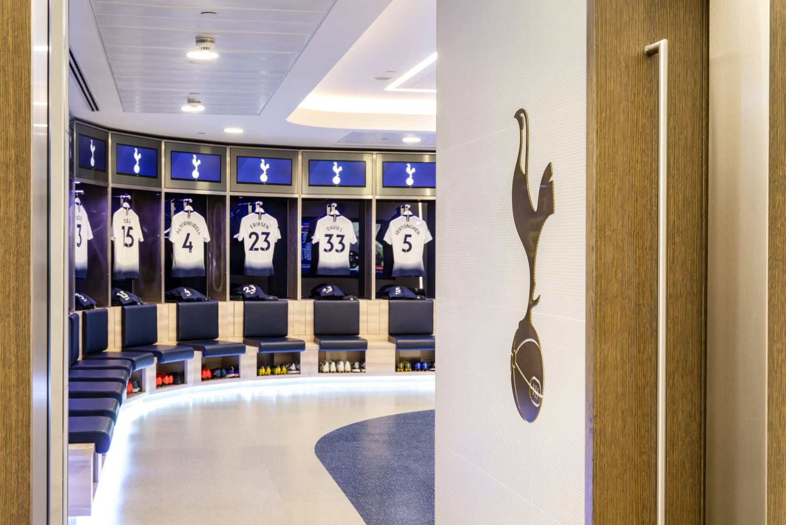 A modern football locker with sport jerseys hung up above the blue seating. Spotlights illuminate the empty space, creating a sense of anticipation and professionalism.