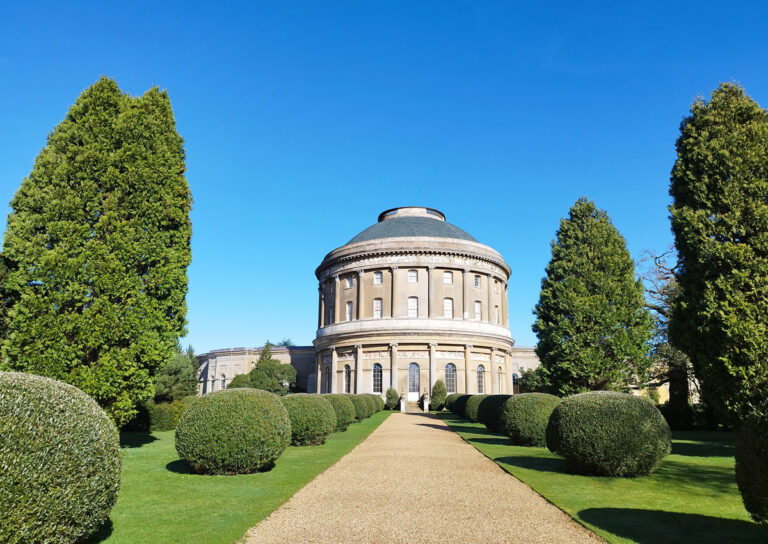 A grand neoclassical building with a large dome, surrounded by hedges and tall trees, under a clear blue sky, creating a serene atmosphere.