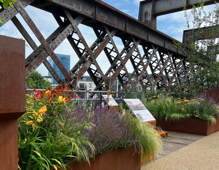 Raised garden beds with colourful and bright flowers and grasses which line a wooden walkway.