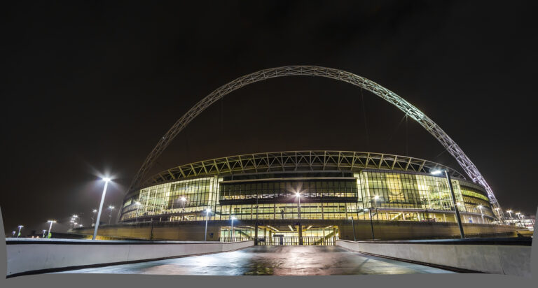 A tied-arch bridge illuminated with lights at night, set against a dark sky, with the city skyline visible in the background.