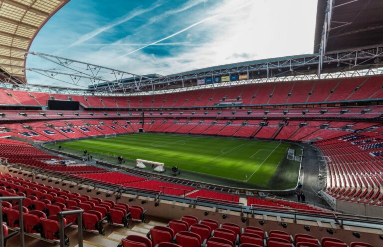 A large sports stadium featuring a vibrant green field surrounded by seating. The sky above is partly cloudy, and the structure has a red exterior.