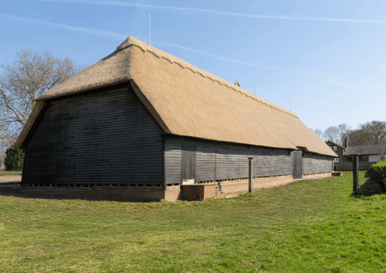 A large barn with a thatched roof and wooden stalls, on a grassy area under blue skies.
