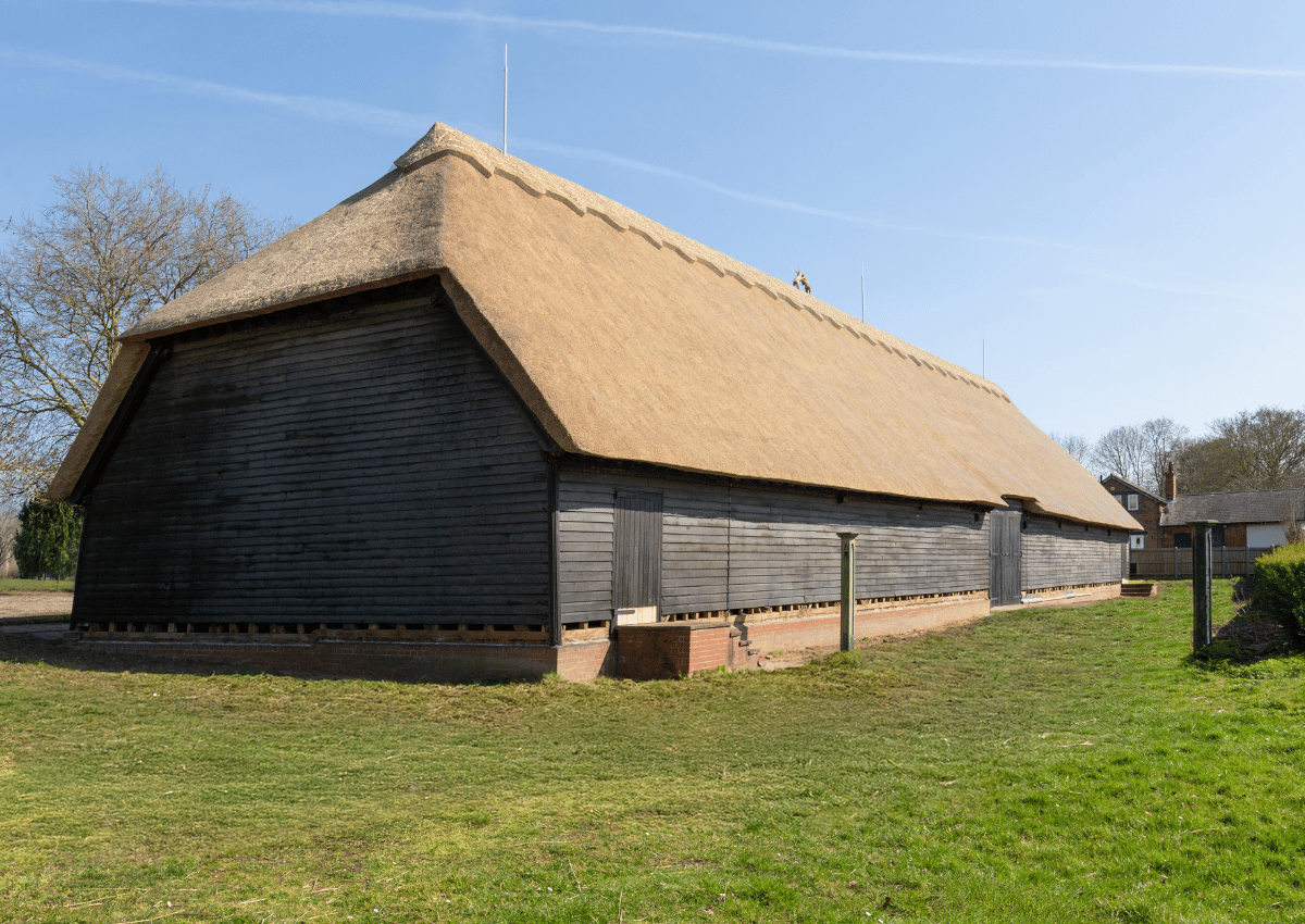 A large barn with a thatched roof and wooden stalls, on a grassy area under blue skies.