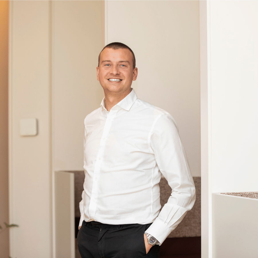 Man smiling, wearing a white shirt and dark trousers, standing indoors with one hand in pocket near a light-coloured wall.