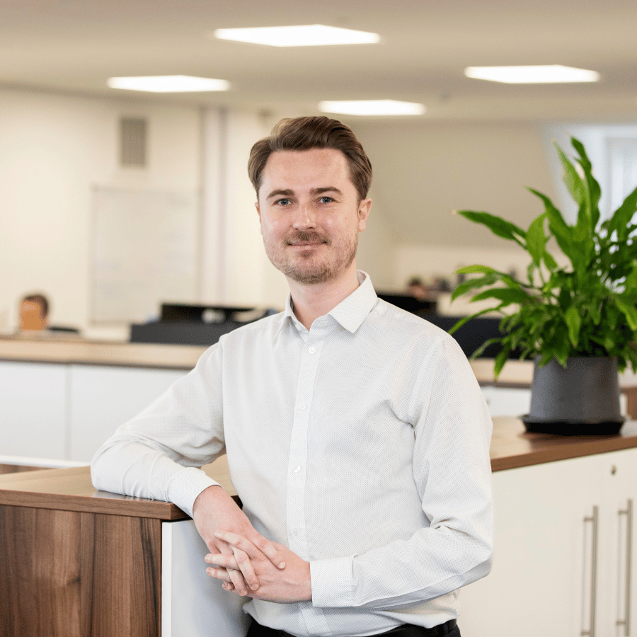 Man wearing a light-coloured shirt, standing in a modern office setting with a wooden counter and a potted plant.