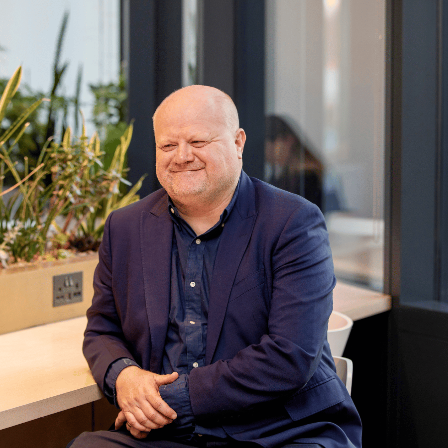 Man in a navy suit seated at a modern indoor workspace with plants on a counter and large windows in the background, smiling.