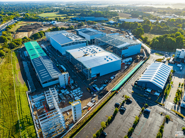 Aerial view of a large construction site with multiple buildings in various stages of completion, surrounded by greenery and a parking lot with cars.