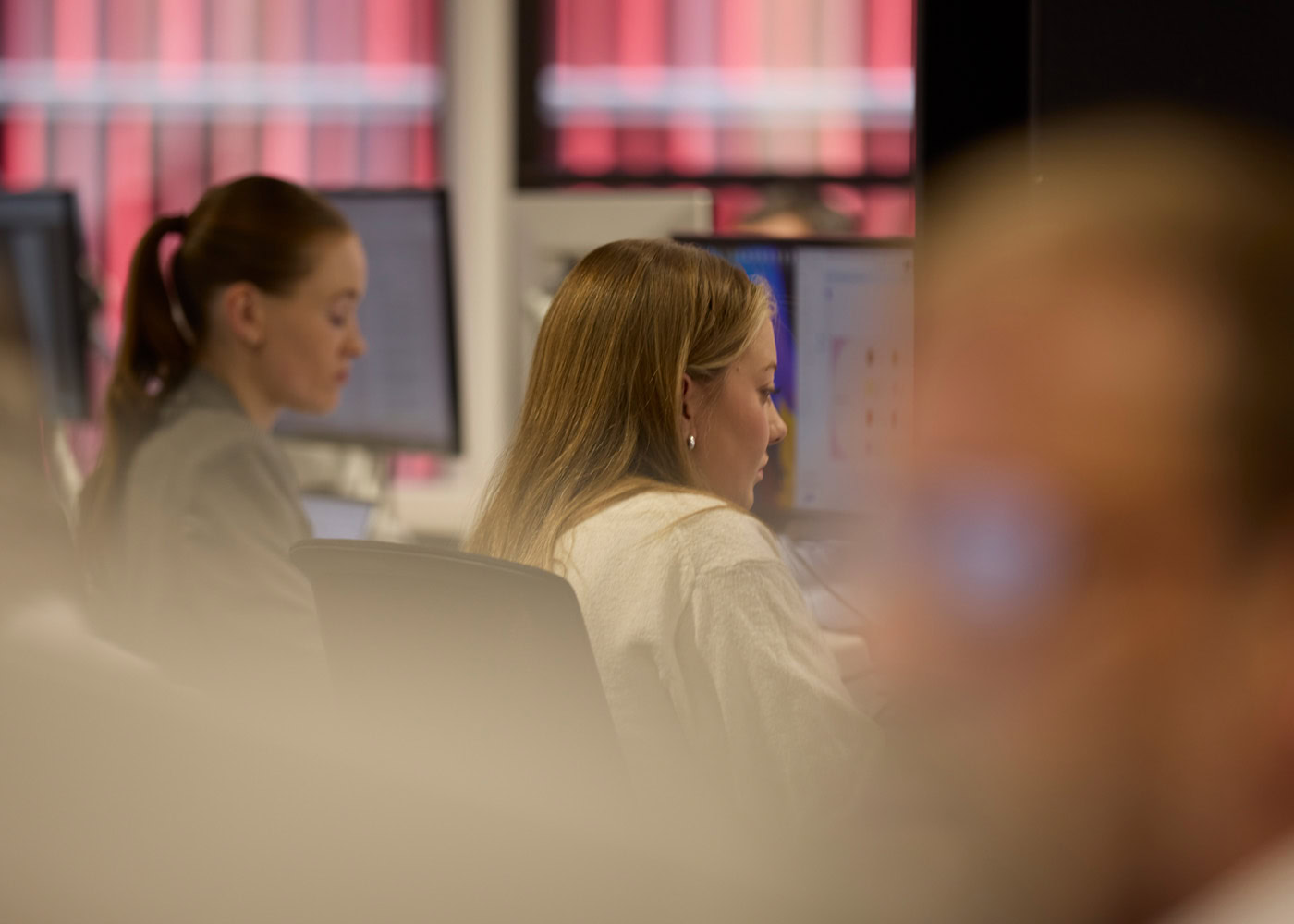 Two woman sitting next to each other in the office on their computer screens with blurred people in the background, creating a concentrated mood.