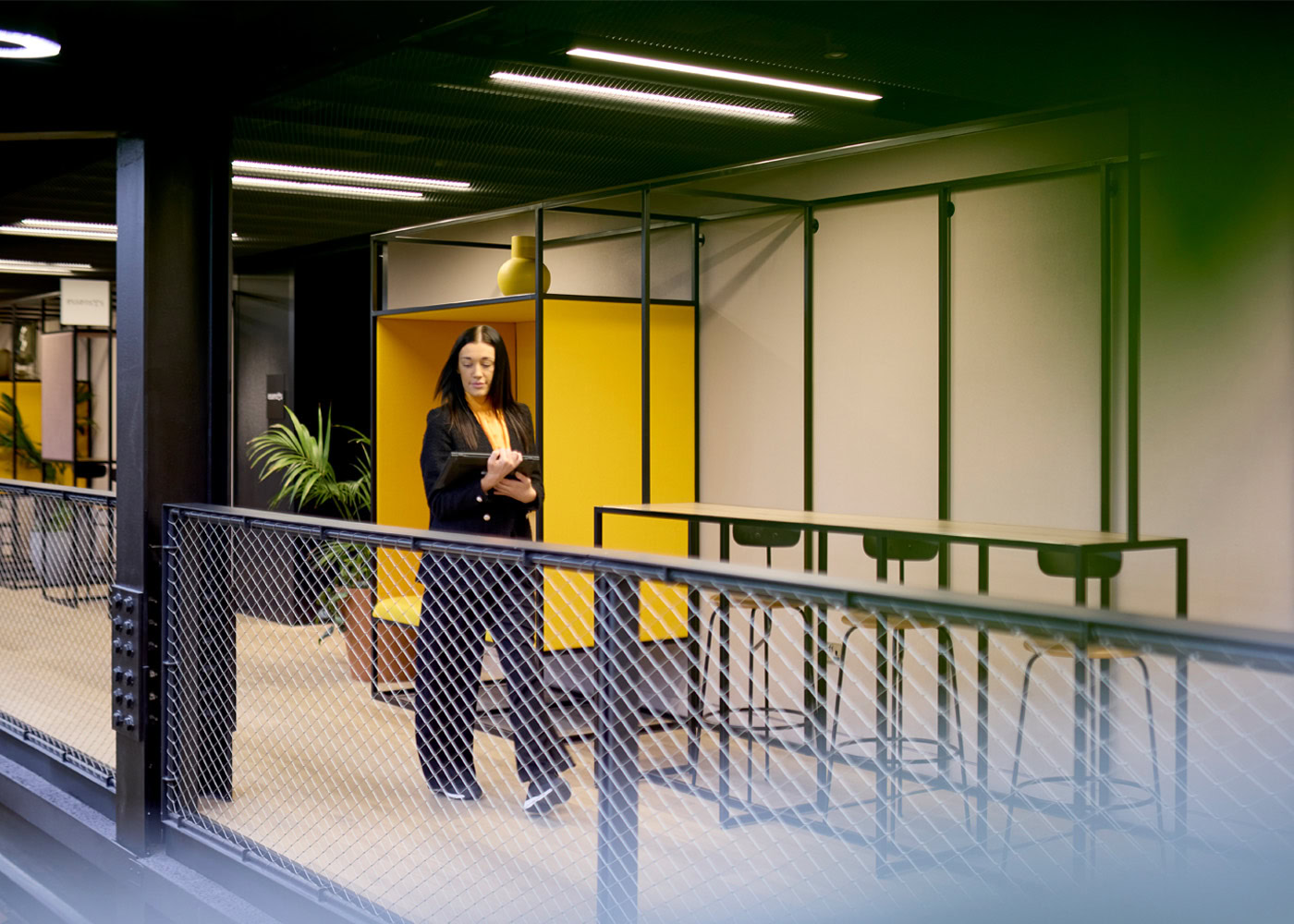 A woman standing in an office space carrying a tablet. There are tall tables, stool and some green plants in the background.
