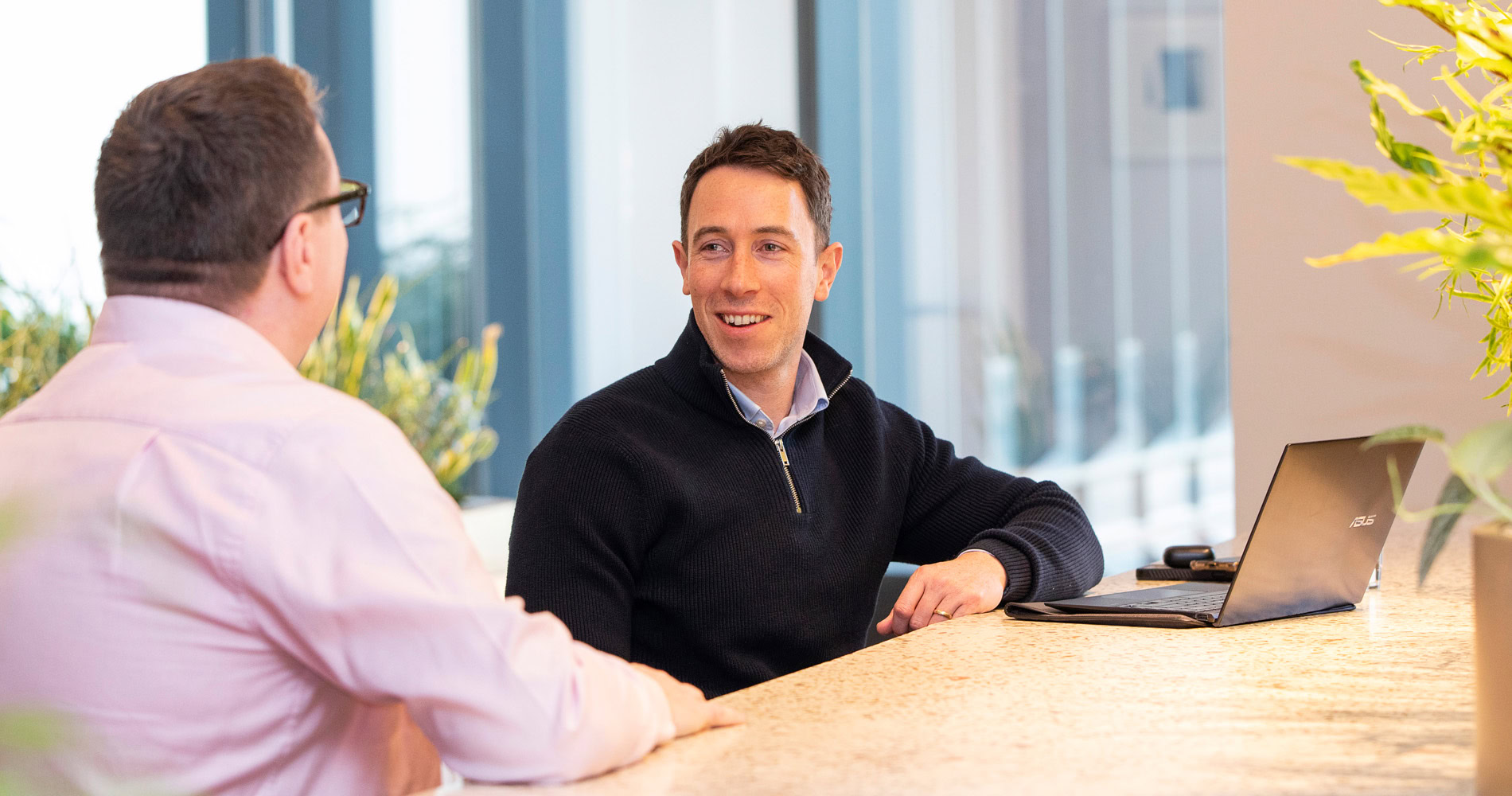 Two men sitting on a bright, office table, having a conversation. One has a laptop open, creating a friendly, collaborative and professional atmosphere