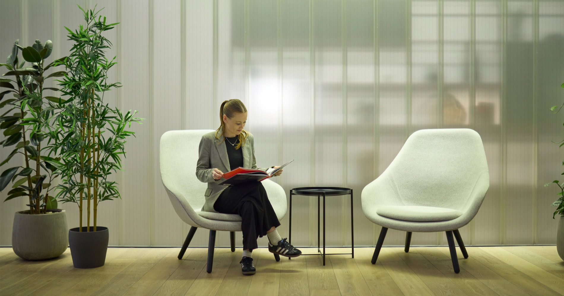 A woman reading a book on a white chair. Next to her is two potted plants with another empty chair the other side.