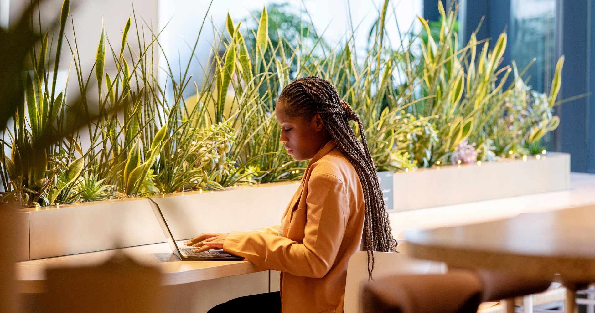 A woman in a yellow blazer typing away on a laptop. She is surrounded by tall green plants.