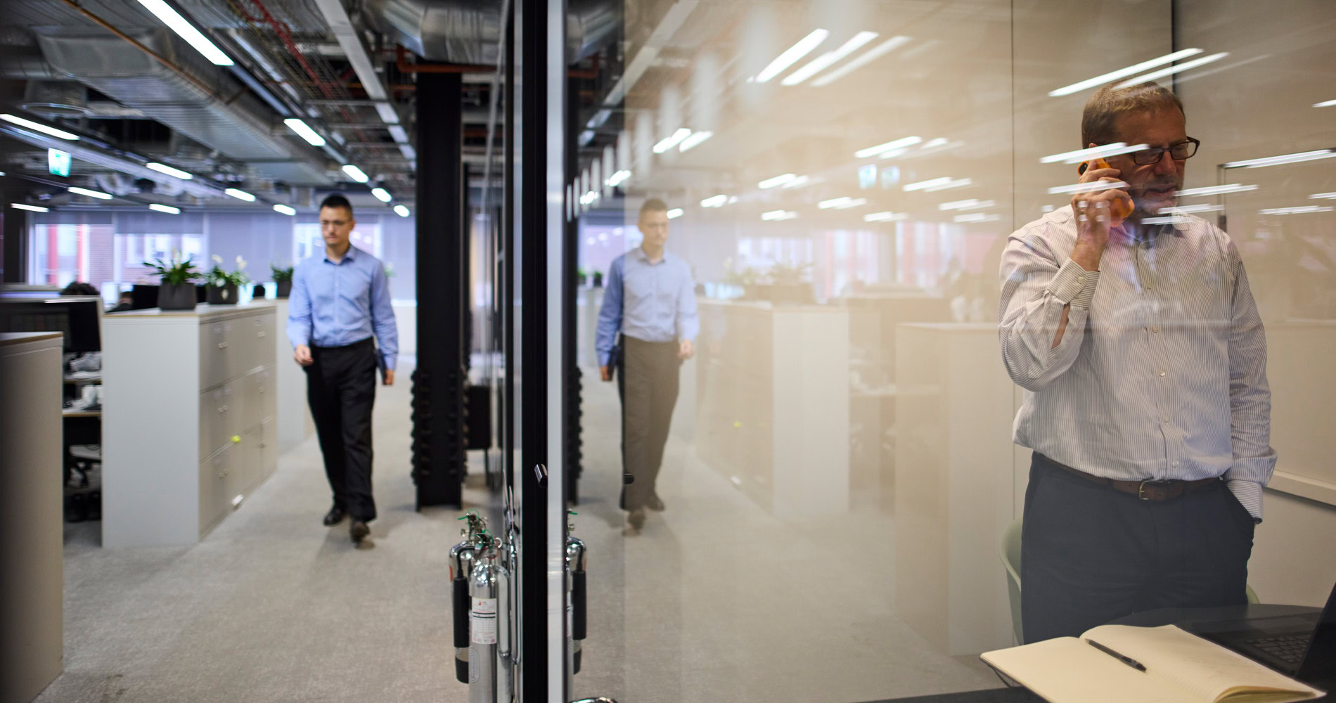 A man in a blue shirt walking in an office corridor with reflections on the glass wall; with another man talking on the phone.