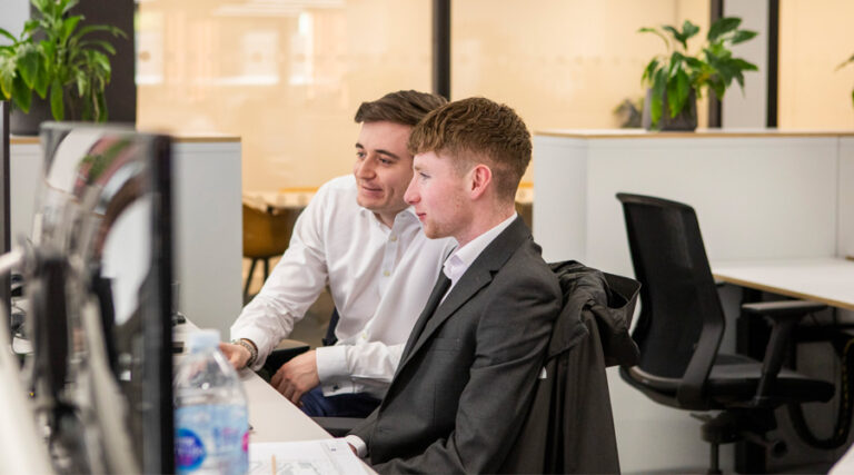Two men in the office looking at a monitor. One is wearing a white shirt, the other in a grey suit. Two green plants in the background, creating a calm atmosphere surrounding them.