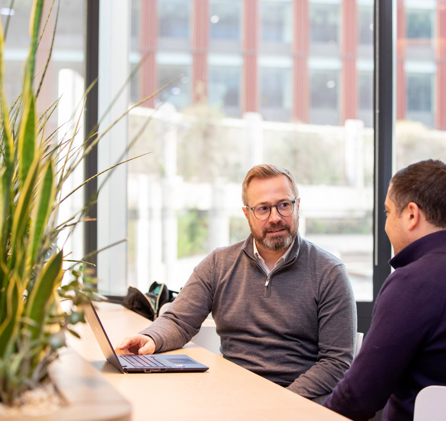 Two men sitting with a laptop, having a conversation. Featuring a large window and indoor plants.