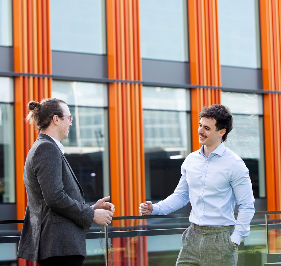 Two men chatting and smiling on a glass balcony. In the background there is a modern building with orange panels.